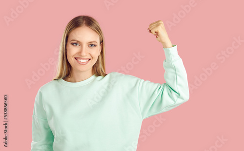 Woman power. Beautiful smiling woman raises her hand showing her biceps as sign of her strength and confidence. Portrait of caucasian blonde woman with make-up and sweatshirt on pink background.