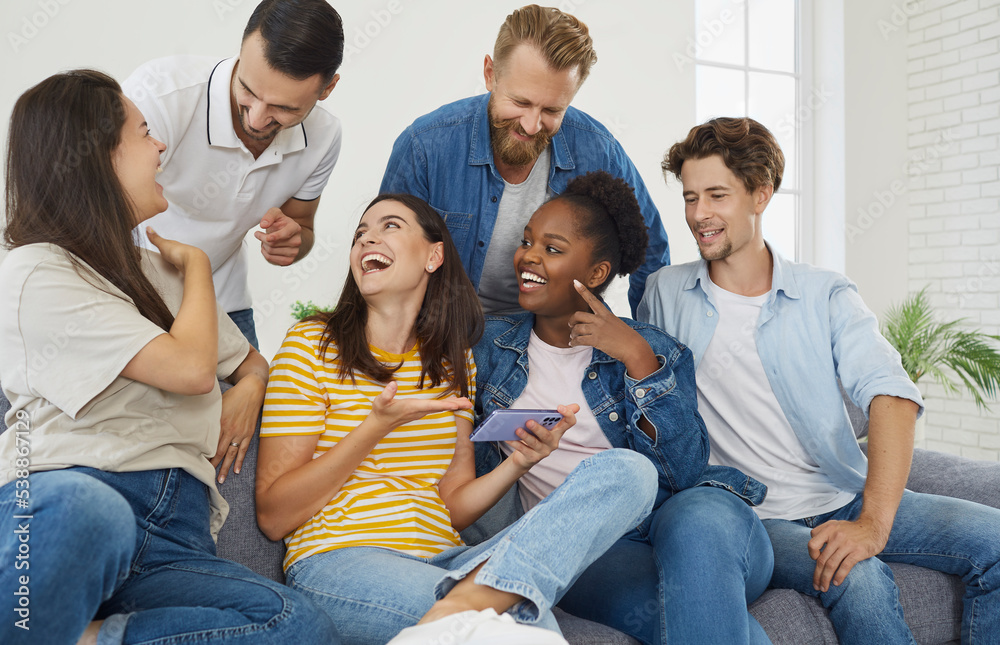 Friendship. Cheerful company of friends watching funny videos on smartphone screen during friendly gatherings at home. Multiracial man and woman talking laughing sitting on sofa in living room. Friendship. Cheerful company of friends watching funny videos on smartphone screen during friendly gatherings at home. Multiracial man and woman talking laughing sitting on sofa in living room.