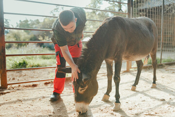 Caretaker with down syndrome taking care of animals in zoo, stroking donkey. Concept of integration people with disabilities into society.