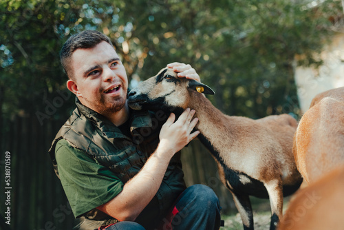 Caretaker with down syndrome taking care of animals in zoo, stroking goat. Concept of integration people with disabilities into society. photo