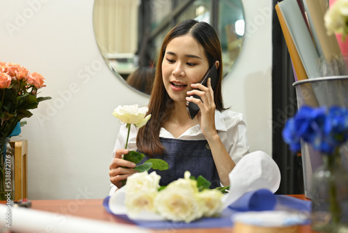 Pretty young woman talking on mobile phone, taking orders from clients while sitting in her floral shop