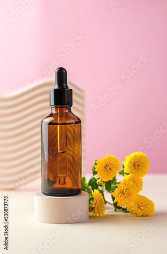 Essential oil or serum and yellow chrysanthemum flowers on a beige podium. Amber bottle with pipette.
