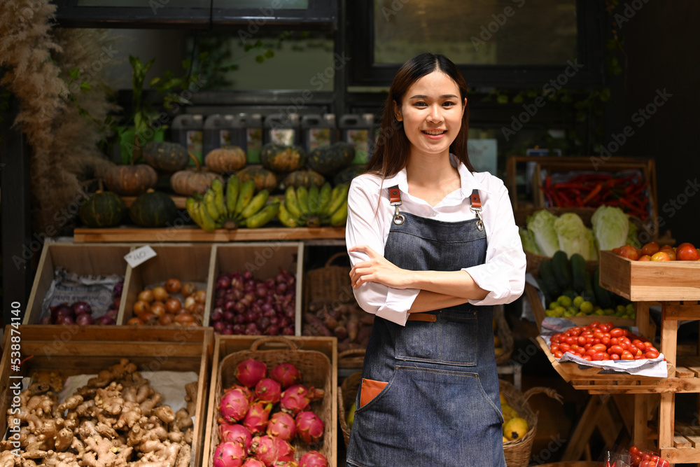 Portrait of friendly woman owner in apron standing with crossed arms front of organic fruit and vegetable stalls at a farmers market