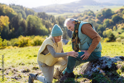 Photography Senior man with hurting knee resting during autumn walk, his wife taking care of him