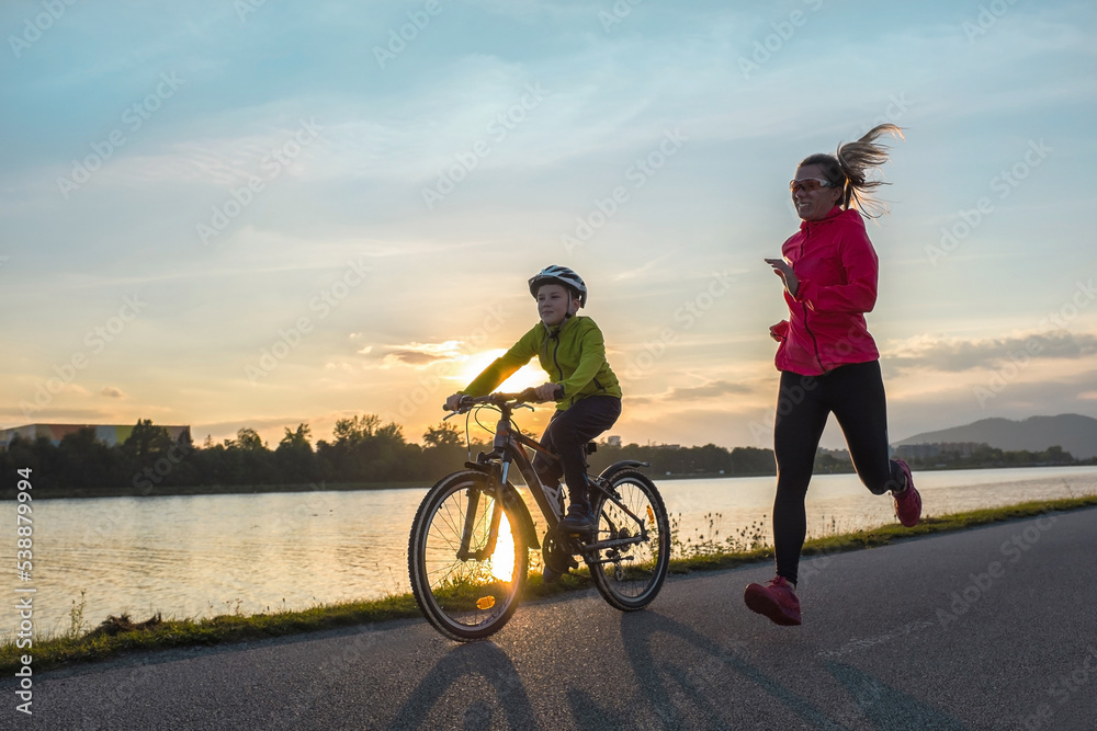 Happy mother and son go in sports outdoors. Boy rides bike in helmets, mom runs on sunny day. Silhouette family at sunset. Fresh air. Health care, authenticity, sense of balance and calmness