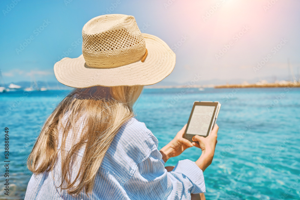 Fototapeta premium Fashion pretty woman outdoors lifestyle watching, reading on tablet ebook on the beach in summer day. Wearing wide brimmed hat, Sunbating with uv protection. Concept of beach vacation.