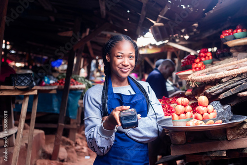 African millennial lady wearing colourful Apron selling tomatoes and vegetables in a typical local african market holding a POS terminal photo