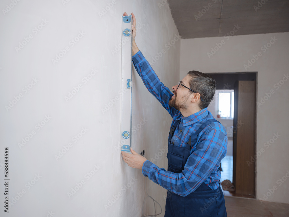 young man in blue work suit doing Repair apartment. Home renovation concept. Cropped shot of handsome bearded young man holding level tool during home improvement young man in blue work suit doing Repair apartment. Home renovation concept. Cropped shot of handsome bearded young man holding level tool during home improvement