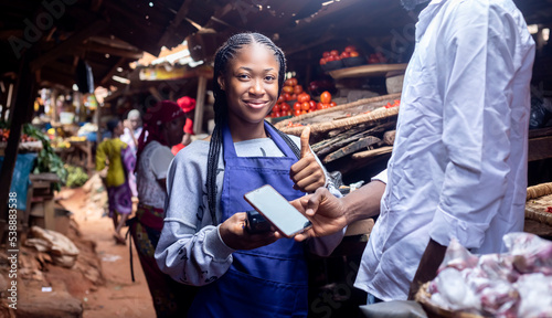 A young African friendly saleswoman passing pos terminal over counter to customer paying with smartphone Smiling given a thumbs up