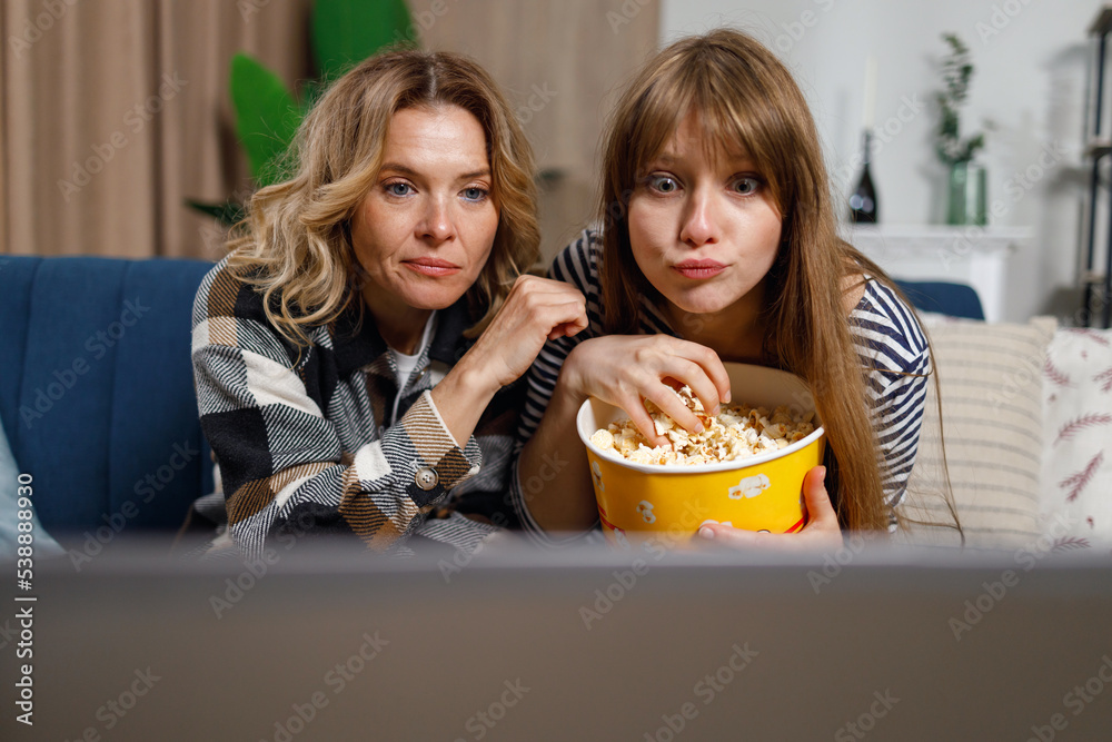 Mature mother and adult daughter watching movie on laptop and eating popcorn indoor Mature mother and adult daughter watching movie on laptop and eating popcorn indoor