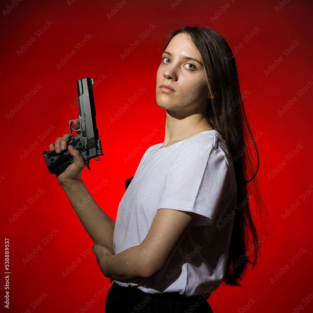 young woman with long hair in a white t-shirt holds a gun in her hand on a red background young woman with long hair in a white t-shirt holds a gun in her hand on a red background
