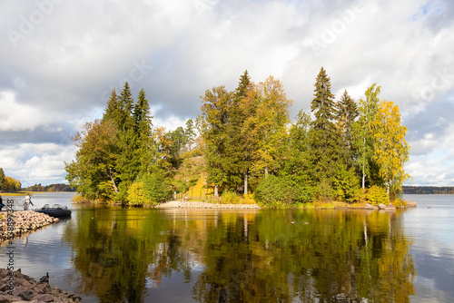 Autumn landscape, trees on lake shore under blue sky with white clouds and people with motorboat