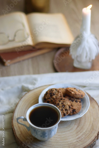Cup of tea or coffee, plate of chocolate chip cookies, open book, reading glasses, candle and vase with flowers. Selective focus.