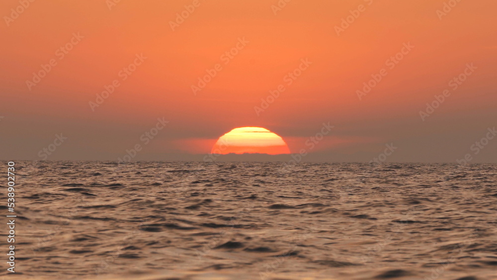 A fire red sunset with the sun behind the clouds falling into the sea by an island in the ocean