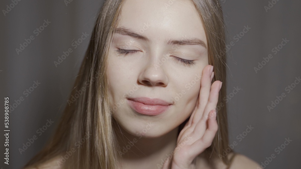 Close-up beauty portrait of young woman with smooth healthy skin, she gently touches her face with her fingers on light grey background and smiles Close-up beauty portrait of young woman with smooth healthy skin, she gently touches her face with her fingers on light grey background and smiles