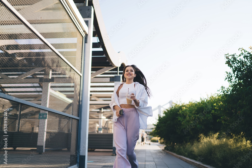 Smiling woman walking on sidewalk with mobile phone near modern construction