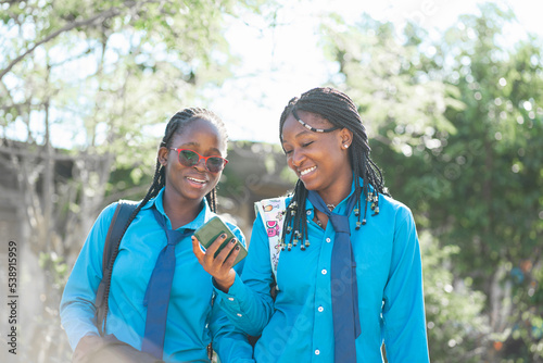 Due giovane studentesse africane in uniforme scolastica guardano contenuti divertenti sullo smartphone mentre vanno a scuola. photo