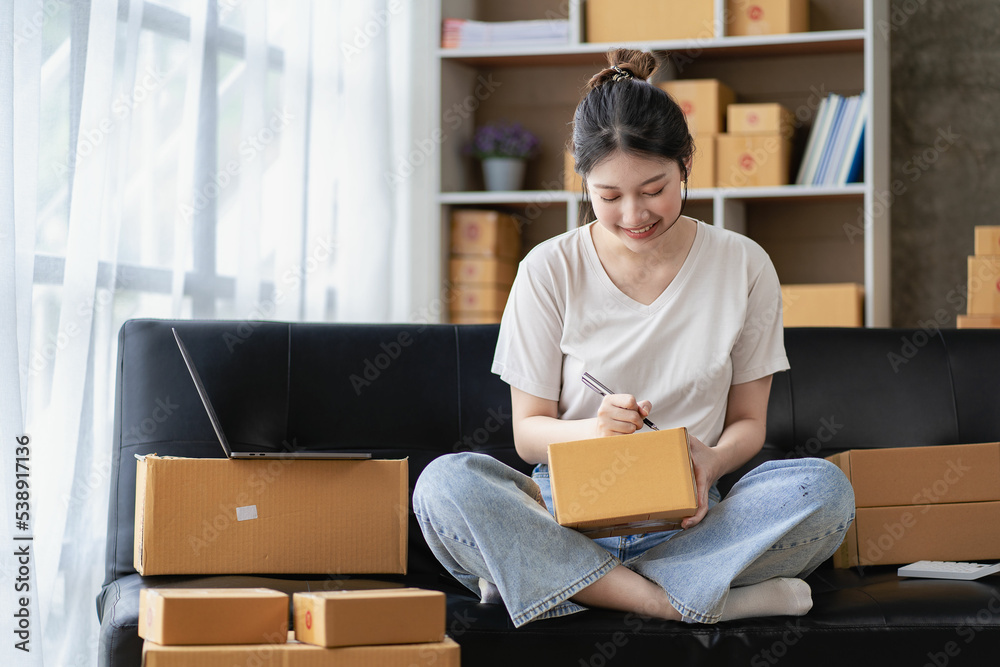 Asian woman sitting on sofa at home working freelance using laptop with box, successful Asian woman with raised hand, online marketing packaging and delivery box, SME concept Asian woman sitting on sofa at home working freelance using laptop with box, successful Asian woman with raised hand, online marketing packaging and delivery box, SME concept