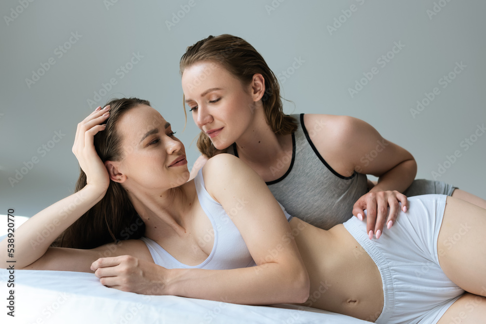 young and sexy lesbian couple smiling at each other while lying on bed in underwear isolated on grey. young and sexy lesbian couple smiling at each other while lying on bed in underwear isolated on grey.