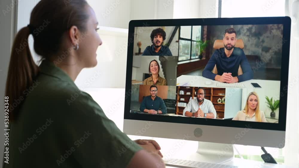 Young freelancer woman greeting participants of online meeting, has video conference with colleagues on the distance, view over shoulder woman using computer app for video call to diverse team