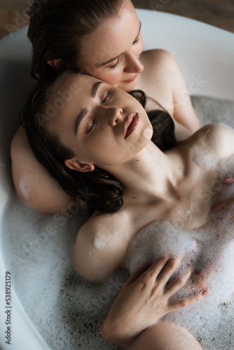 top view of lesbian woman hugging girlfriend in bathtub with closed eyes.