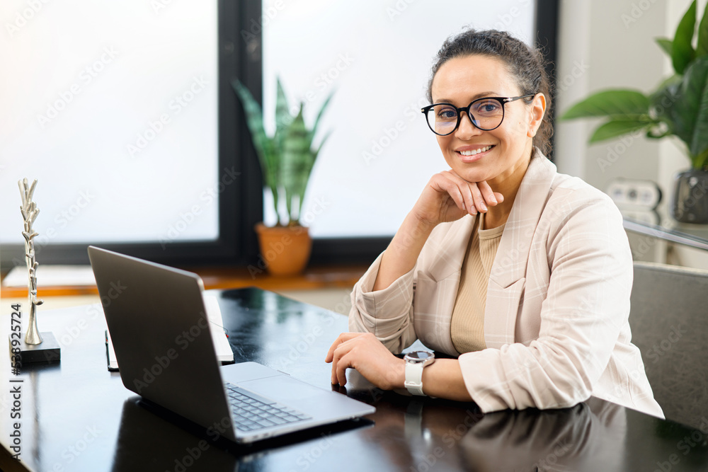 Confident middle-aged woman work with a laptop at the modern office, female employee in smart casual wear looks at the camera and smiles, typing messages, answering email, front view Confident middle-aged woman work with a laptop at the modern office, female employee in smart casual wear looks at the camera and smiles, typing messages, answering email, front view
