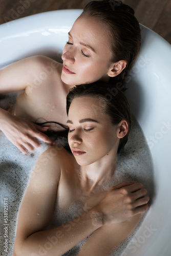 top view of naked lesbian women relaxing in bubble bath with closed eyes.