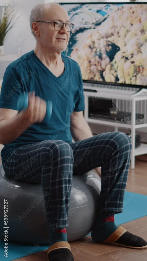 Vertical video: Senior man lifting dumbbells and sitting on fitness toning ball to do physical training and exercise. Retired person doing workout activity with weights to work arms muscles at home. Vertical video: Senior man lifting dumbbells and sitting on fitness toning ball to do physical training and exercise. Retired person doing workout activity with weights to work arms muscles at home.