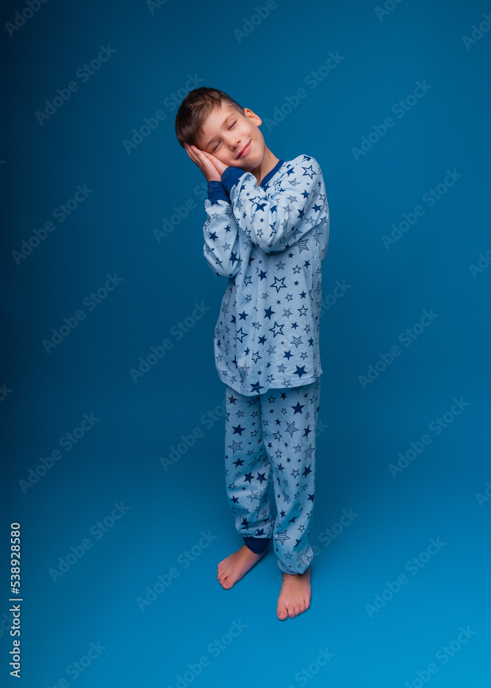Portrait of a cute little child standing with his hands closed, pretending to sleep isolated on a blue background. a boy in pajamas Portrait of a cute little child standing with his hands closed, pretending to sleep isolated on a blue background. a boy in pajamas