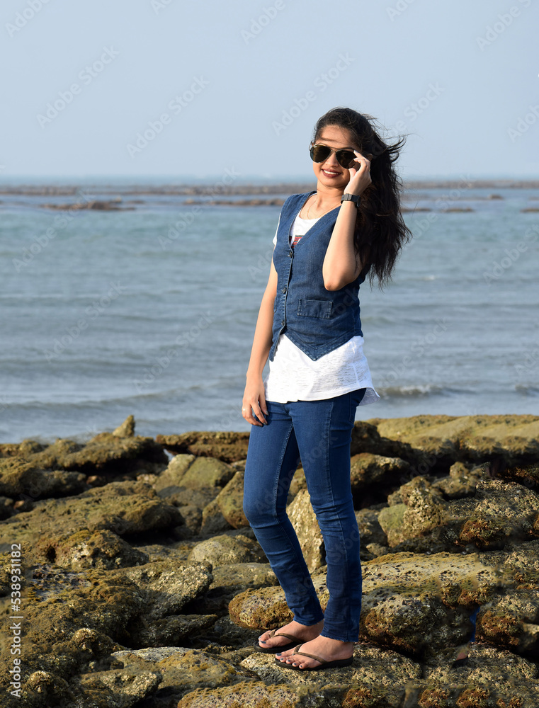 Stylish girl standing on the coral of an ocean enjoying the nature. Young woman holding the sun glass standing at sea beach
