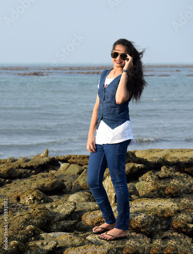 Stylish girl standing on the coral of an ocean enjoying the nature. Young woman holding the sun glass standing at sea beach