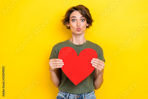 Portrait of adorable funny girl brown hair wear khaki t-shirt hold large paper heart pouted lips isolated on yellow color background