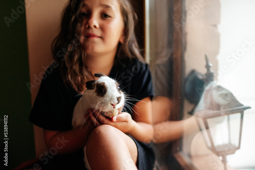 a girl sits on the window and holds a white rabbit with brown ears on her knees