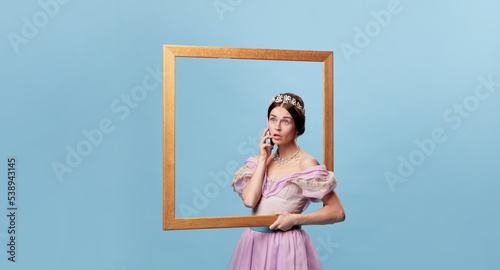 Emotional girl, actress wearing medieval princess or countess dress standing with picture frame over blue background. Portrait through picture frame.