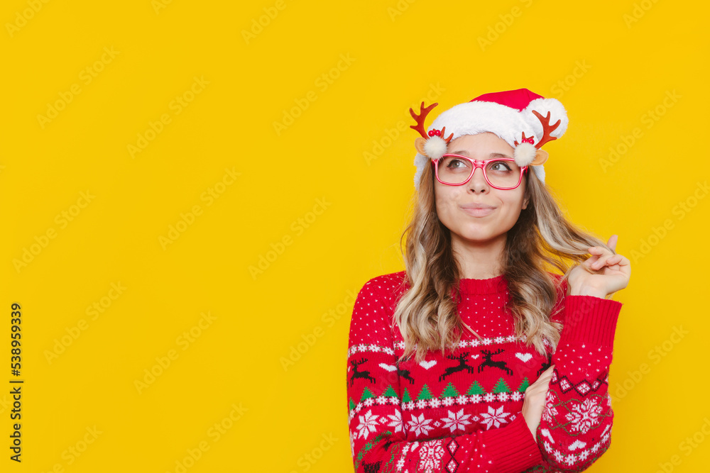 Young thoughtful blonde woman in glasses with deer antlers, a red warm sweater and Christmas Santa hat thinks about something looks away at empty copy space isolated on a color yellow background Young thoughtful blonde woman in glasses with deer antlers, a red warm sweater and Christmas Santa hat thinks about something looks away at empty copy space isolated on a color yellow background