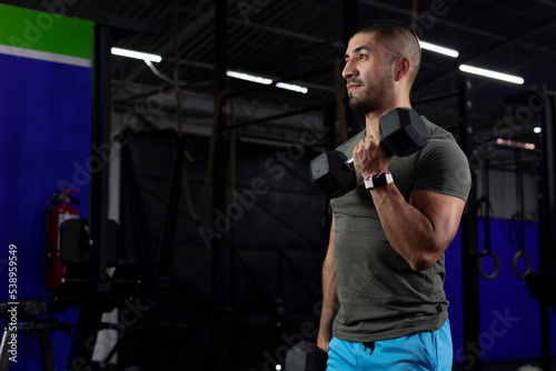 latino man with sportswear doing biceps exercises with two dumbbells in a gym