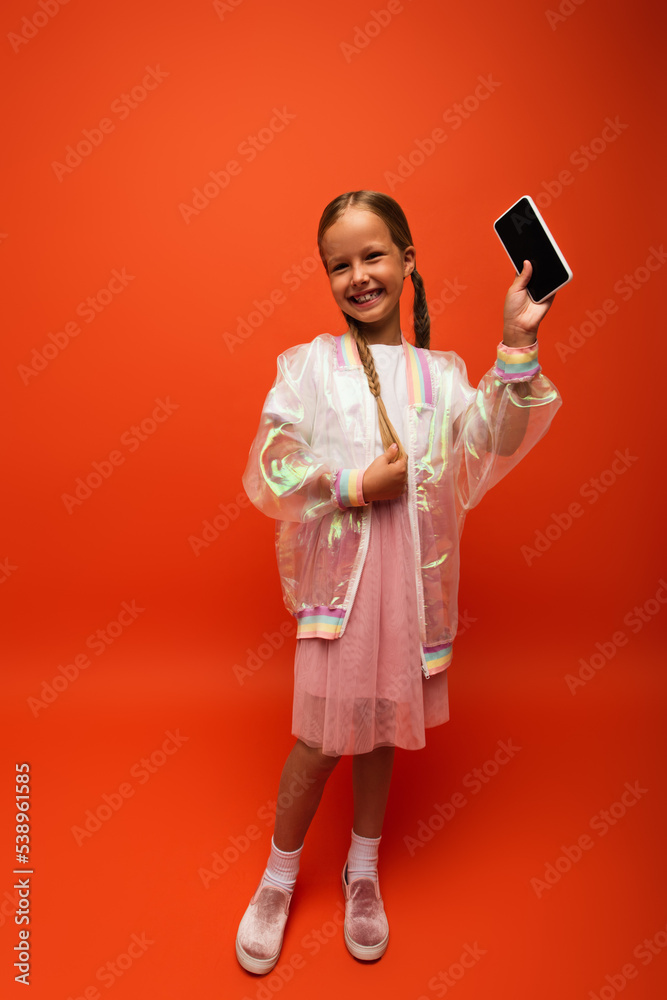 full length of cheerful girl touching pigtail and showing smartphone with blank screen on orange background
