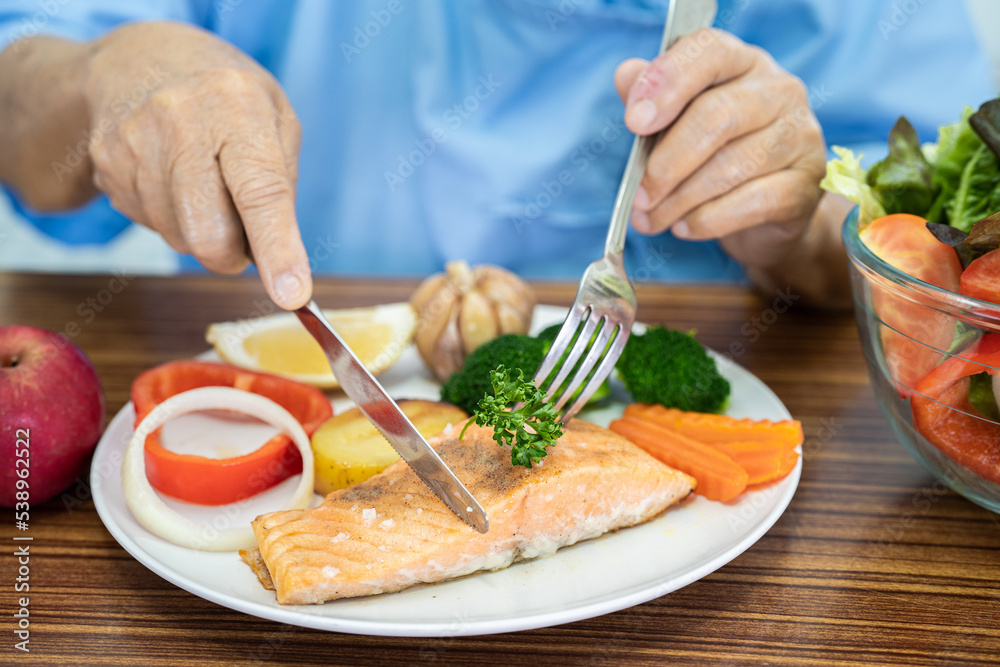 Asian senior or elderly old lady woman patient eating Salmon steak breakfast with vegetable healthy food while sitting and hungry on bed in hospital.