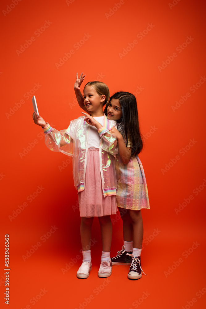 full length of happy girls waving hand and pointing with finger while taking selfie on orange background full length of happy girls waving hand and pointing with finger while taking selfie on orange background