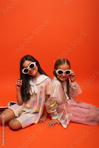 smiling preteen girls in trendy clothes and sunglasses looking at camera while sitting on orange background