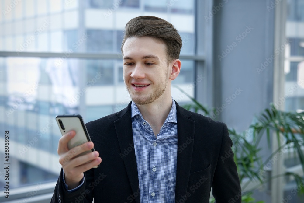 Portrait of happy handsome guy, young European blonde man in shirt is using his cell mobile phone, looking at screen of smartphone, reading or typing message and smiling in office building indoors. Portrait of happy handsome guy, young European blonde man in shirt is using his cell mobile phone, looking at screen of smartphone, reading or typing message and smiling in office building indoors.