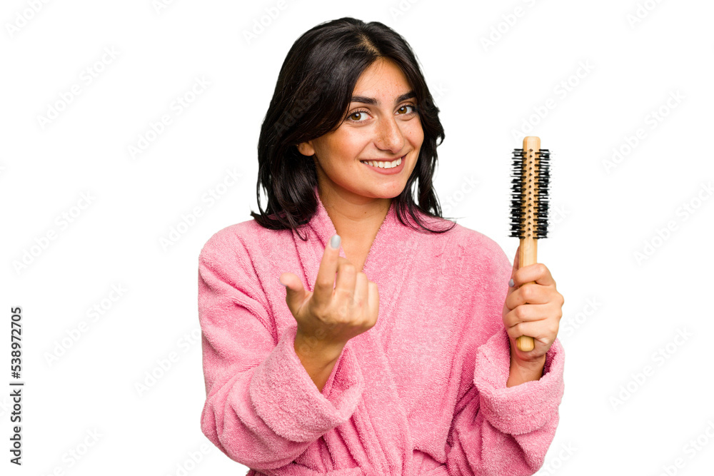 Young Indian woman in a bathrobe holding an hairbrush isolated pointing with finger at you as if inviting come closer. Young Indian woman in a bathrobe holding an hairbrush isolated pointing with finger at you as if inviting come closer.