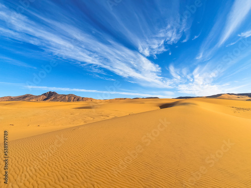 Textures sand ripples in foreground and a rocky mountain with a blue sky and stratus clouds with motion effect. Colorful smartphone photography in Tadrart Rouge, Djanet Algeria. Tassili N'Ajjer Sahara