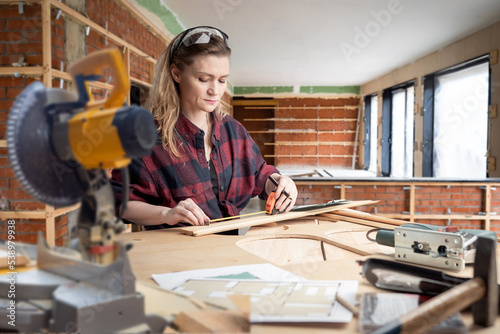 Woman furniture designer. Girl measures piece of wood with tape measure. Woman design wood furniture. Girl carpenter in house under construction. Woman works as carpenter. Lady furniture designer
