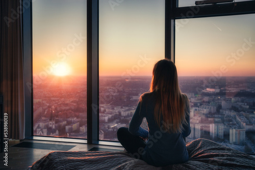 A young woman sitting on a bed and watching the sunset through the windows from a high floor, luxurious apartment with a view on the city.