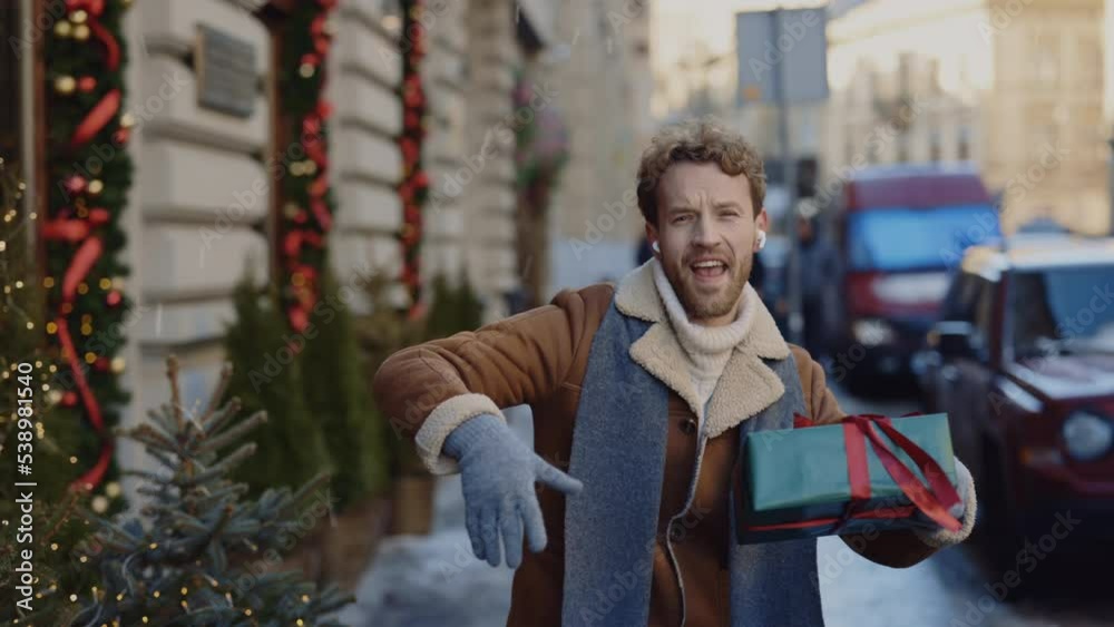 Caucasian crazy man going dancing on the decorated street listening to music using earphones singing holding the christmas gift. People and christmas holidays concept