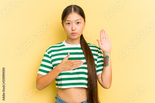 Fototapet Young Asian woman isolated on yellow background taking an oath, putting hand on chest
