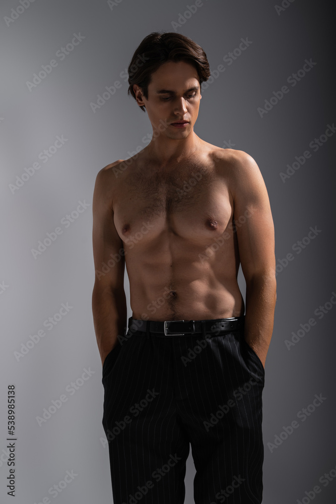 good looking man with muscular body posing with hands in pockets of black trousers on grey