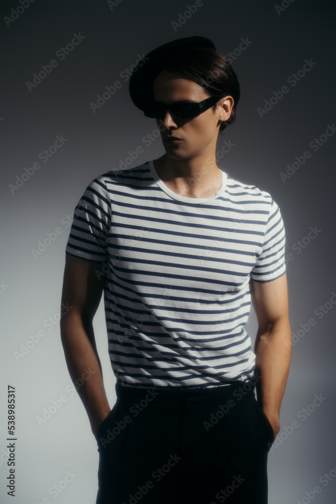 Stylish man in beret and sunglasses holding hands in pockets of shorts on grey background with lighting Stylish man in beret and sunglasses holding hands in pockets of shorts on grey background with lighting