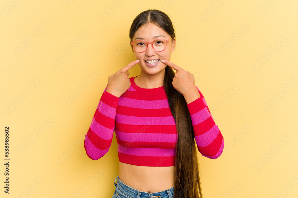 Young Asian woman isolated on yellow background smiles, pointing fingers at mouth. Young Asian woman isolated on yellow background smiles, pointing fingers at mouth.